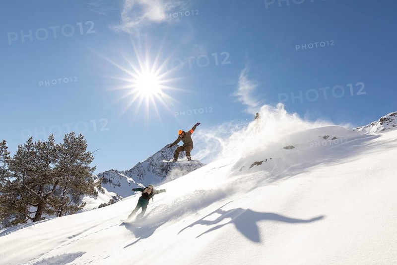 Two snowboarders carve through fresh powder in a stunning backcountry setting under a clear blue sky. The image captures the thrill of freeride snowboarding as the sun shines brightly
