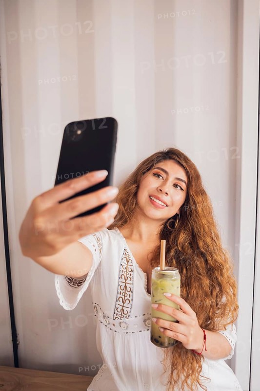 A young American Latin woman enjoys her lifestyle by taking a selfie with her smartphone while holding a trendy bubble tea drink, showcasing a relaxed and joyful moment