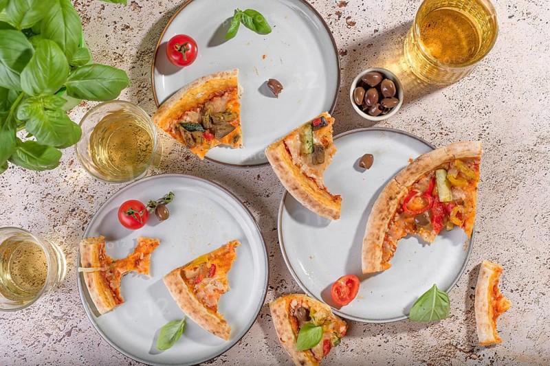 Top view of rustic vegetarian pizza slices with zucchini, eggplant, bell peppers, and basil. Accompanied by olives and refreshing drinks on a rustic tabletop setting