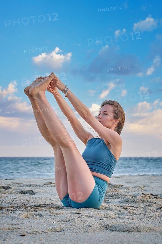 A woman balances gracefully in Ubhaya Padangusthasana on sandy beach. The sunset adds a warm glow to the scene, showcasing her flexibility and strength in this challenging yoga pose