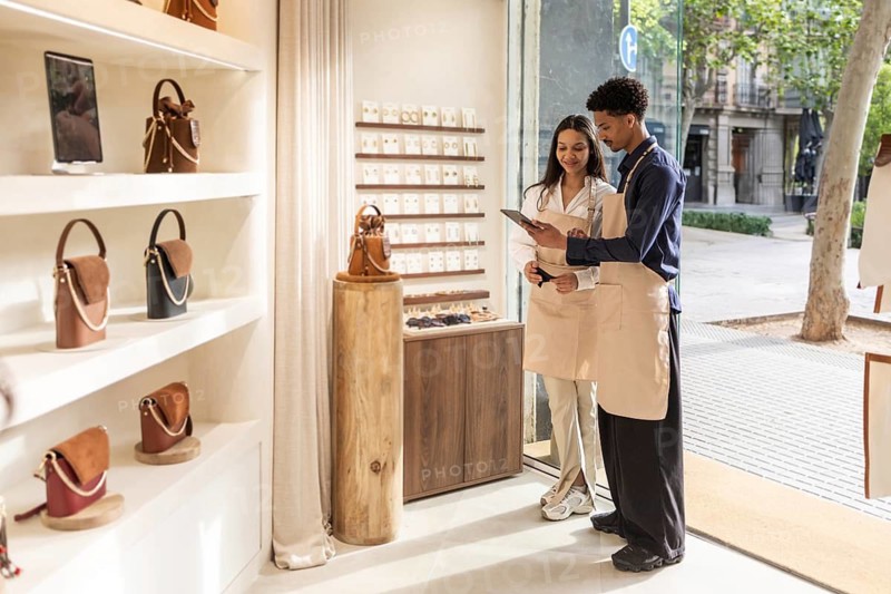 Two employees, clad in aprons, examine a digital tablet while standing in a chic bag store with natural light. Shelves display various handbags in a modern setting