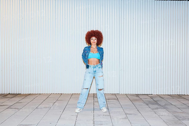 African descendent woman with voluminous afro hair posing confidently in front of a ribbed metal wall. She wears stylish ripped jeans, a blue crop top, and a denim jacket, complemented by round sunglasses