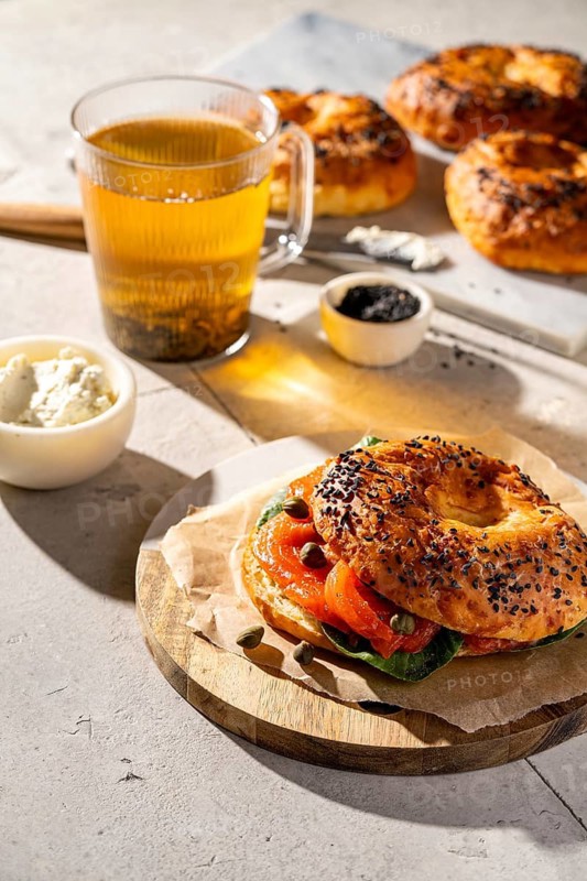 A close-up of a sesame seed bagel with smoked salmon, cream cheese, and capers. Accompanied by a glass of tea, this breakfast setup offers a savory delight