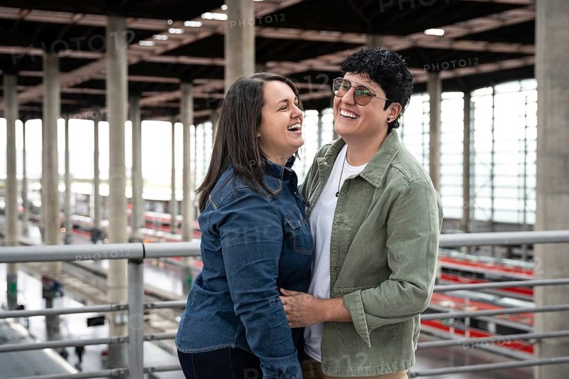 A joyful lesbian couple shares a moment of laughter in a train station. They are embracing each other, highlighting love and happiness amidst city life