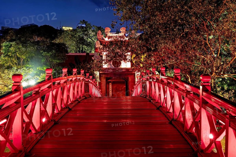 A red-lit bridge leads to a temple surrounded by trees in a mystical night-time atmosphere, The Ngoc Son Temple in Hoan Kiem Lake in Hanoi Vietnam