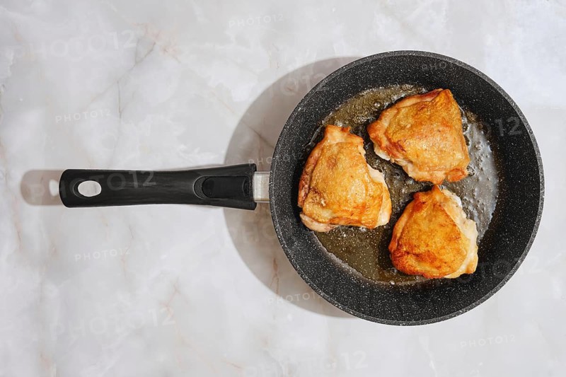 Three chicken thighs are sizzling in a frying pan on a marble countertop. The cooking process shows the browning of the meat. This scene takes place in a kitchen during the day