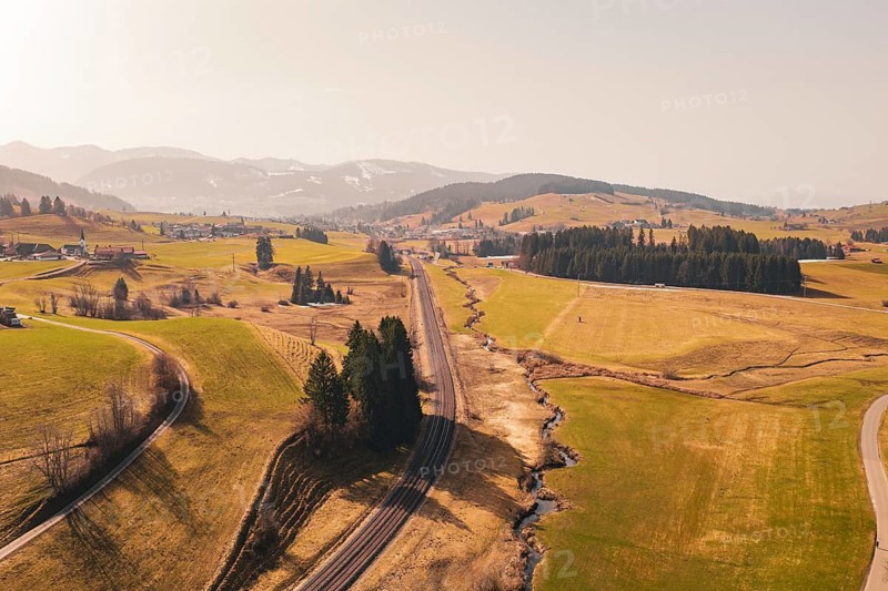 A railway line runs through open fields and hills, framed by trees and mountains, Oberreute, Allgäu, Germany