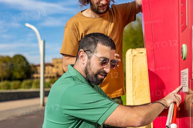 A visually impaired man receives assistance from a friend, highlighting trust and cooperation They interact with a red tactile information board outdoors