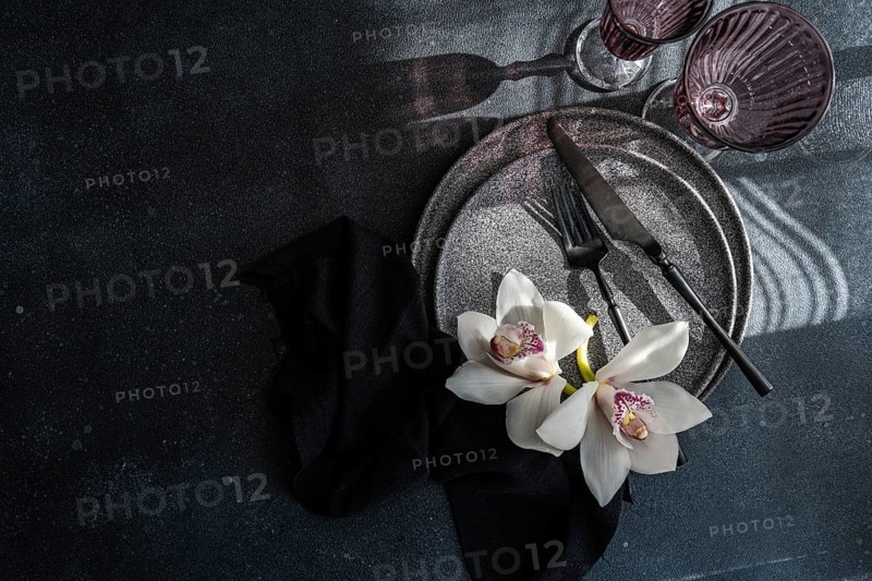 A sophisticated table setting with gray plates, dark cutlery, and delicate white orchids. The scene is bathed in soft light, evoking a sense of elegance and style