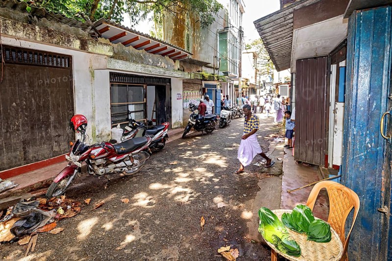Narrow street with leaves on the ground, some motorized vehicles, local architecture in the background, The Gemstone Quarter in the city of Ratnapura in Sri Lanka