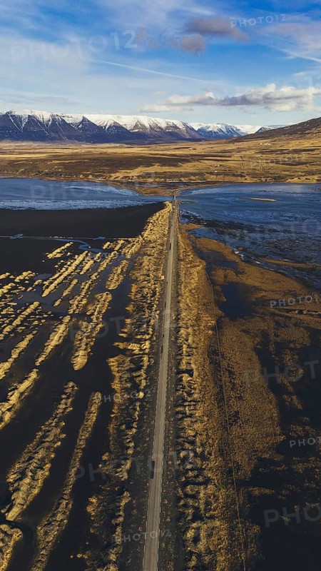 Vertical drone shot of a road stretching through the rugged Icelandic landscape near Hofsos, with distant snow-capped mountains and coastal wetlands under a partly cloudy sky