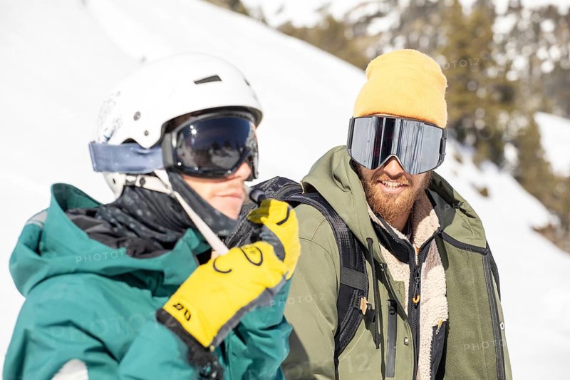 Two snowboarders equipped with gear enjoying a sunny day in the snowy backcountry. They are dressed for cold conditions, wearing helmets and goggles, ready for an adventure