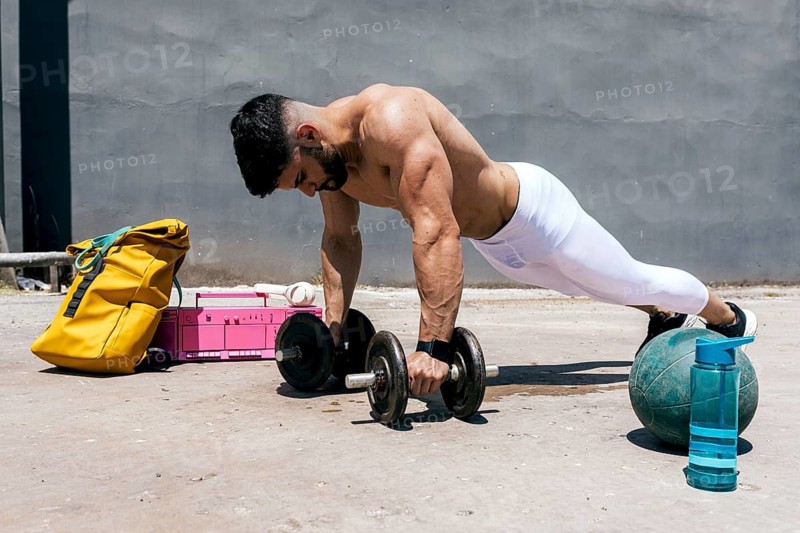 A physically fit man performs an intense outdoor workout using dumbbells and a medicine ball, displaying strength and focus against a concrete backdrop on a sunny day