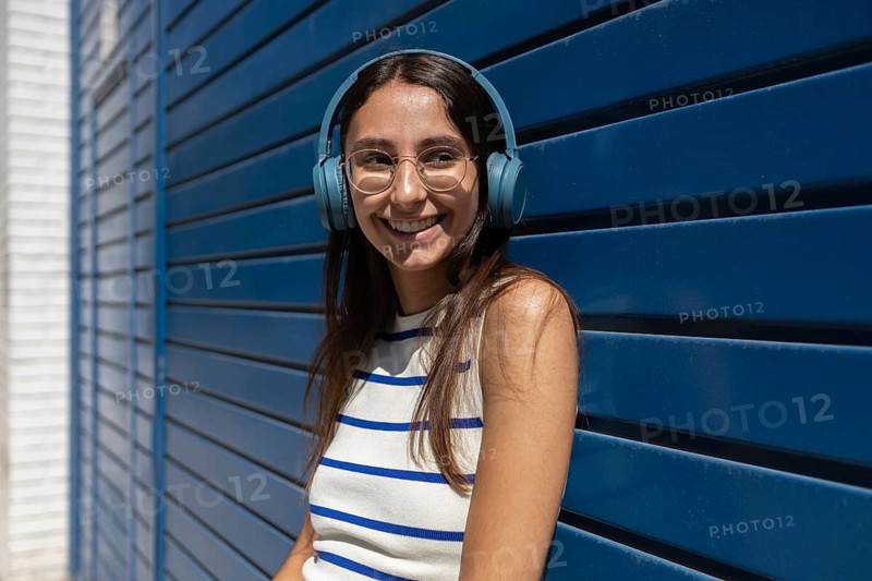 A woman enjoys her music while leaning against a modern blue wall in the city. She embodies a vibrant urban lifestyle, wearing headphones and a stylish striped top