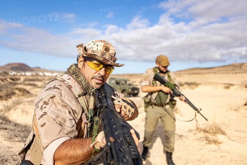 Two soldiers in camouflage gear conduct training in a desert landscape, aiming rifles with intensity. The tactical exercise focuses on combat readiness and strategic movement