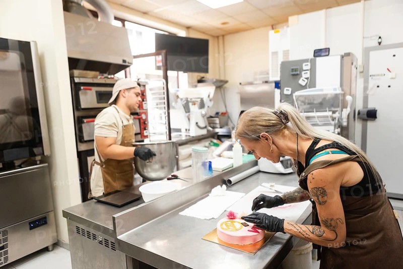 Two skilled pastry chefs work diligently in an Italian bakery, one mixing ingredients and another decorating a cake The bakery setting is lively and well-equipped