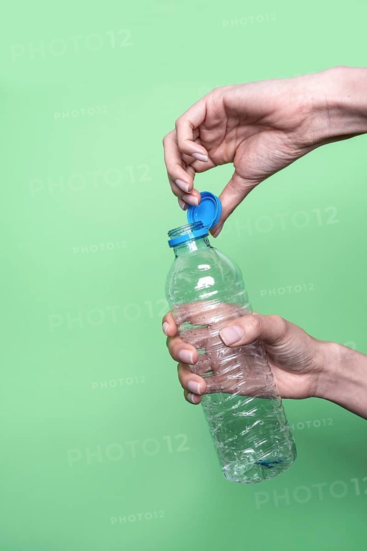 Close-up of hands opening a plastic water bottle, positioned against a vibrant green background. The blue bottle cap contrasts sharply with the green, emphasizing action and color