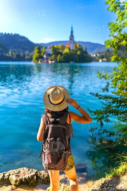 Female tourist wearing backpack and summer hat enjoying breathtaking view of lake bled island with church in julian alps, slovenia
