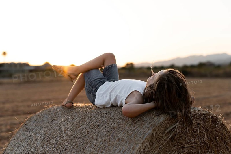 A child in casual summer attire lounges on a hay bale in a rural field as the sun sets, creating a serene ambiance. The image captures the essence of carefree childhood in nature