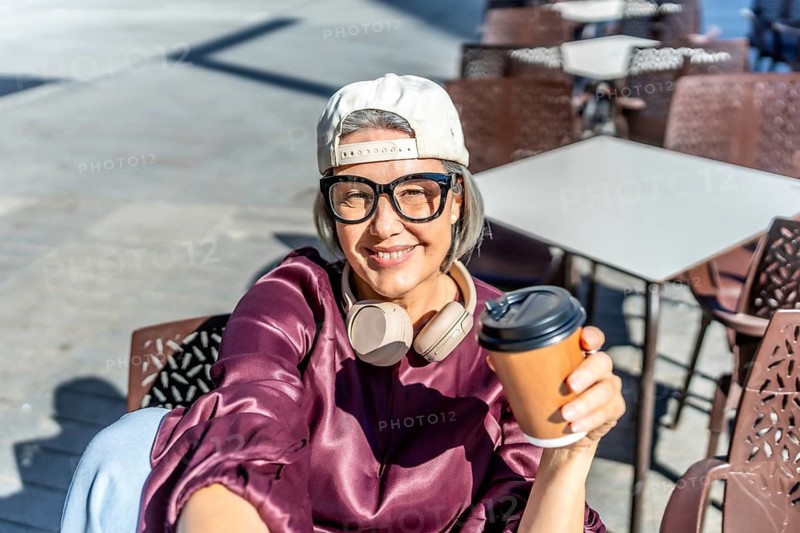 Woman with gray hair smiling at the camera, wearing a cap, glasses, and headphones around her neck, holding a hot coffee cup while relaxing at an outdoor cafe
