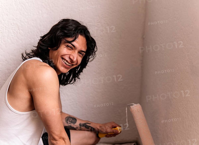Cheerful young man with a brush in hand, painting a white wall in his home, smiling widely. His casual attire and visible tattoos add a personal touch to the scene of home remodeling