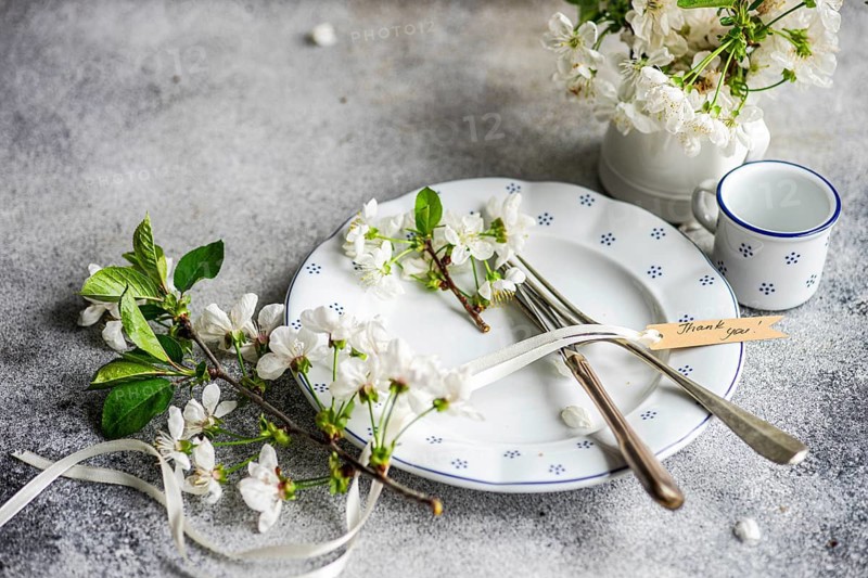 An elegant table setting features a white plate adorned with delicate blossoms and vintage cutlery. A thank you card adds a personal touch to this serene arrangement