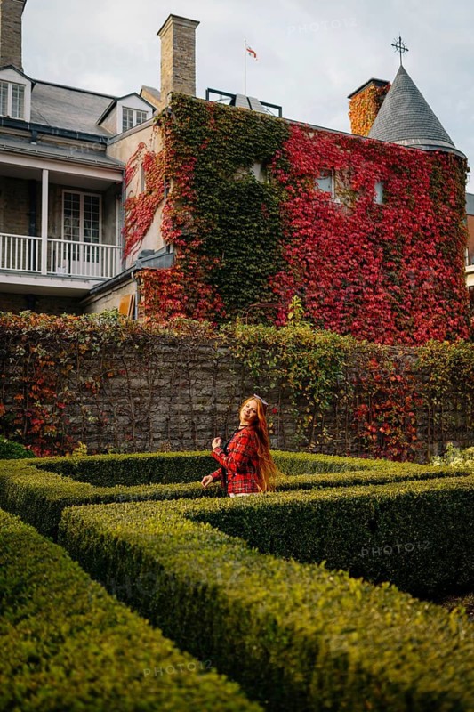 A woman in a red plaid coat stands in a manicured garden at a historic Montreal site, surrounded by vibrant autumn foliage and ivy-covered brick walls