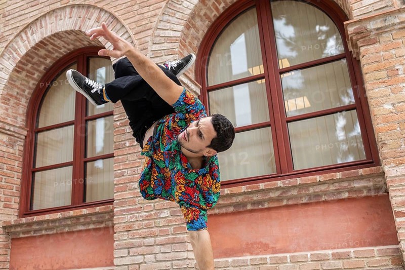 A vibrant Cuban dancer executes an impressive move against a historic brick backdrop. His colorful shirt and dynamic pose capture the essence of lively street dance