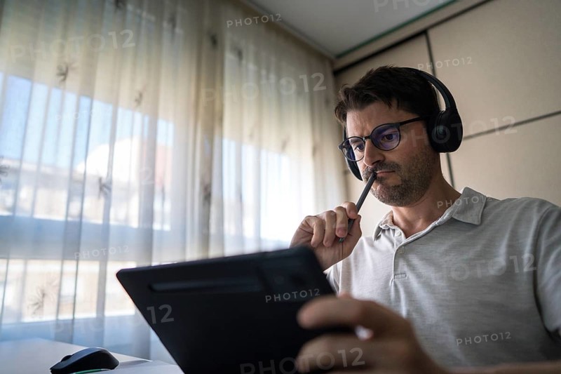 A man sits at home, equipped with headphones, focusing intently on studying Mandarin using a digital tablet, illustrating the modern approach to language learning online
