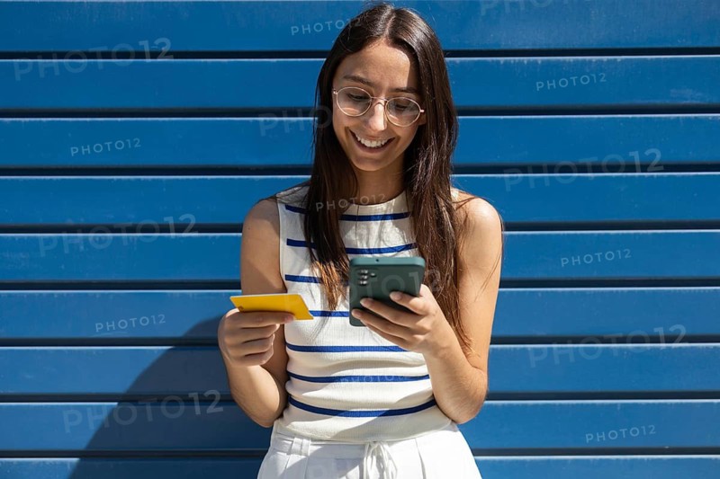 A woman enjoys outdoor city life, engaging with technology through her smartphone and credit card. Her smile reflects a modern, connected lifestyle in an urban setting