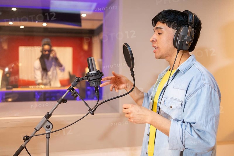 A musician records vocals in a modern studio, wearing headphones and performing passionately into a microphone, while a producer observes through the glass window