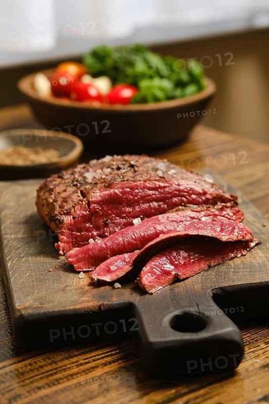 A cutting board holds slices of roasted steak. A bowl of fresh vegetables, including tomatoes and greens, rests in the background. Light illuminates the scene from a nearby window