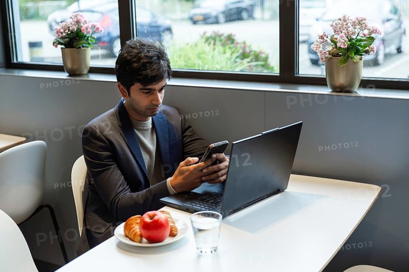 A indian man multitasks efficiently using a smartphone and laptop at a hotel cafe table in amsterdam during a conference of AI. He is enjoying a light breakfast with a croissants and apple