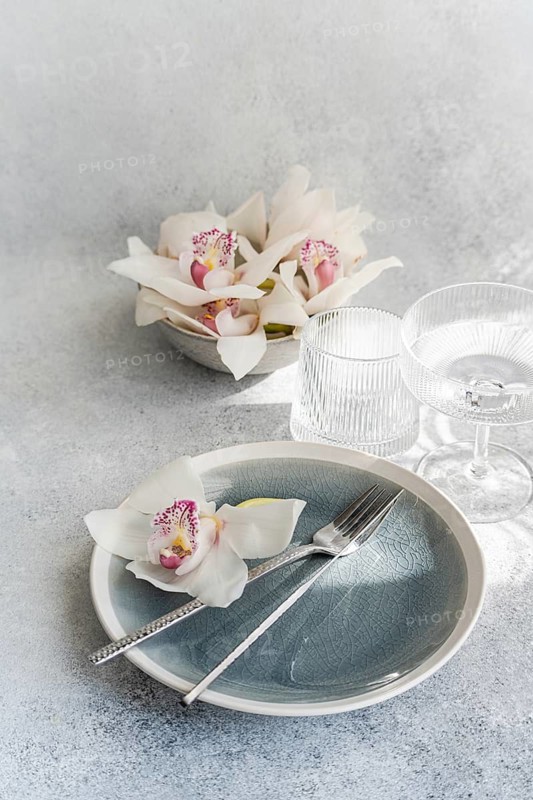 A sophisticated table setting features a blue plate with silver cutlery, adorned with delicate white orchids. Crystal glassware complements the fresh and elegant atmosphere
