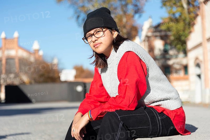 A young Latina in a vibrant red top and black beanie sits in a sunny urban setting Her casual street style and confident expression reflect a modern, youthful vibe