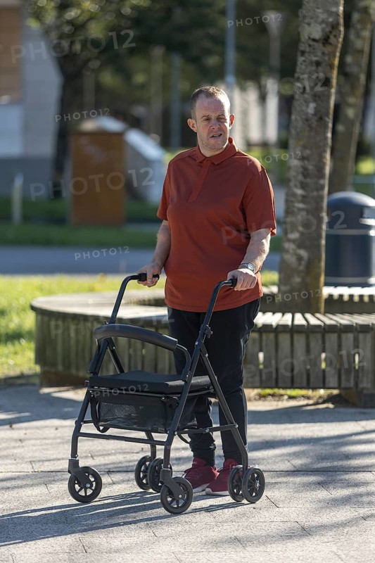 A man with an intellectual disability uses a mobility walker while strolling through a park. His expression reflects determination and independence