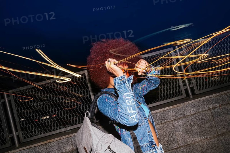 An African descendent woman with vibrant afro hair expresses creativity using light trails in the dark. She captures the motion with her smartphone, creating a dynamic and artistic night scene
