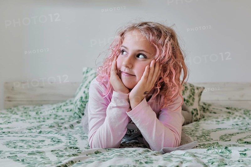 A young girl with pink hair and a dreamy expression rests her head on her hands while lying on a bed with floral patterned bedding, evoking a sense of innocence and warmth