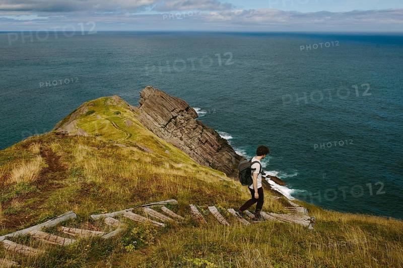 A back view of an unrecognizable male hiker walking down a rugged pathway at Middle Cove in Newfoundland & Labrador, Canada. The lush green landscape and vast ocean create a captivating backdrop