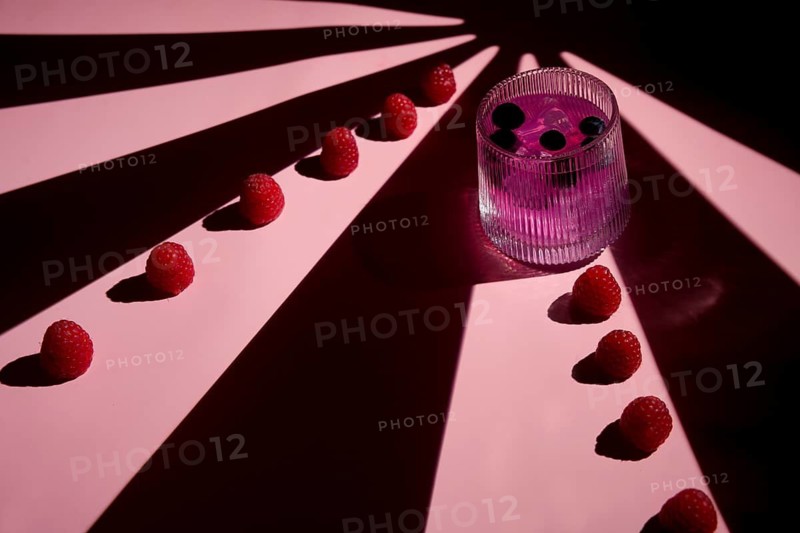 A striking composition featuring a pink drink garnished with berries alongside a row of fresh raspberries. The geometric pink and dark shadows create a vibrant, artistic scene