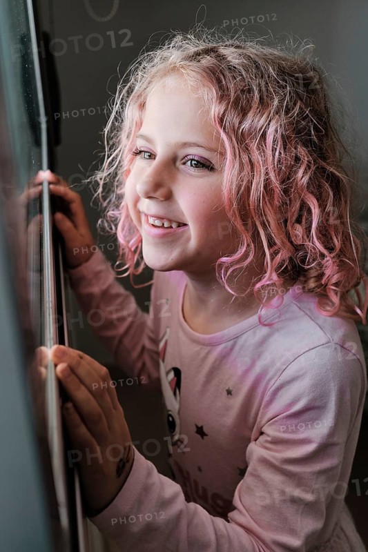 A young child with curly pink hair smiles while peering out a window. The child exudes a sense of wonder and innocence, wearing a cozy pink sweatshirt indoors