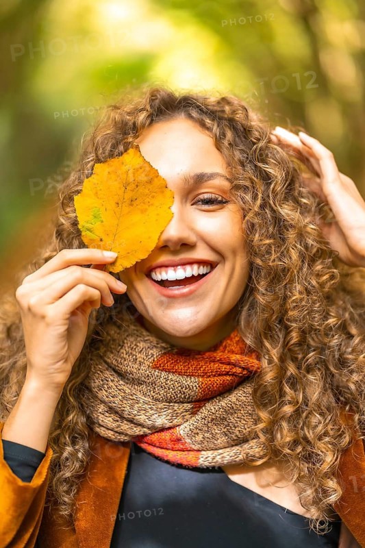 Vertical autumn portrait of a smiling woman with curly hair hiding on a leaf