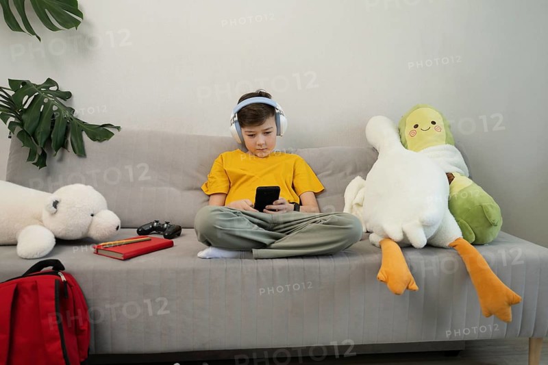 A child in headphones sits on a sofa, engrossed in a smartphone. Surrounded by plush toys and a game controller, the scene depicts modern childhood in a cozy living room