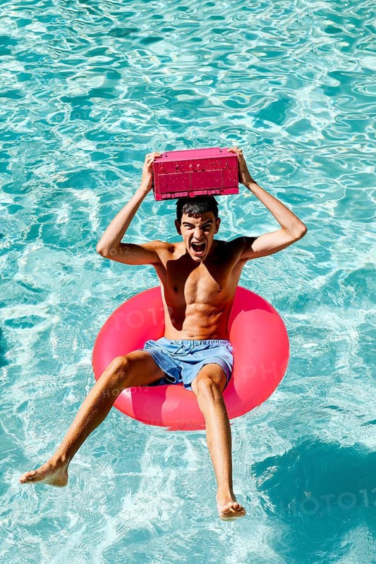 A joyful teenage boy is sitting on a pink float in a pool, holding a pink radio above his head. The bright water reflects the sunny ambiance, evoking summer excitement