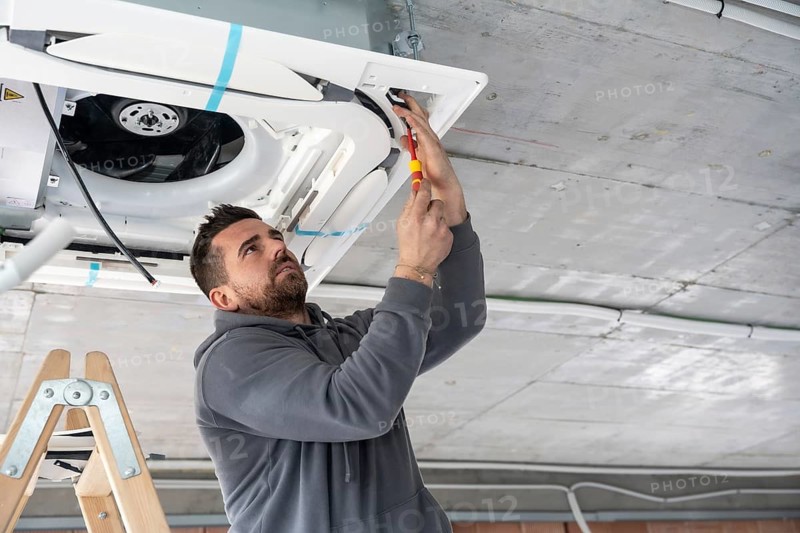 Professional hvac technician standing on a ladder and using a screwdriver to install a new air conditioning unit into a concrete ceiling during construction work