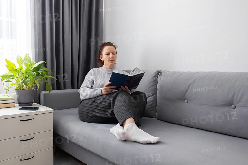 A woman sits comfortably on a gray sofa, reading a book. The serene atmosphere is enhanced by soft lighting, a nearby plant, and a minimalistic interior design