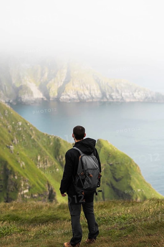 Traveler with backpack stands on a grassy cliff, gazing at a mist-covered fjord and mountains in Norway, capturing the essence of adventure, trekking, and nature