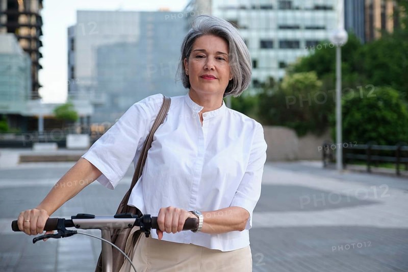 A mature woman with gray hair rides a scooter confidently in an urban environment. She wears a white shirt and appears content, surrounded by modern buildings and greenery