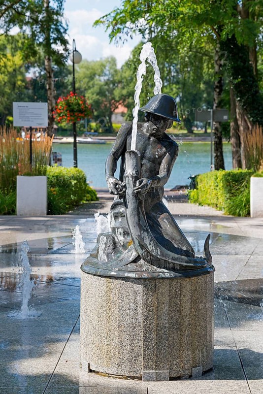 Fountain with statue in a sunny park with trees and water in the background, Ostroda, Osterode in East Prussia, Warminsko-Mazurskie, Warmia-Mazurskie, Poland