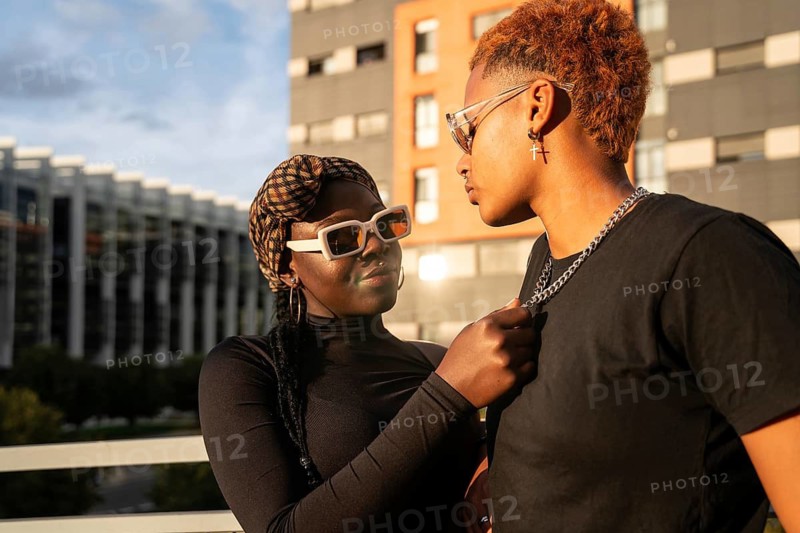A loving LGBT multiethnic couple stands close together in an urban setting as the warm glow of the sunset casts a welcoming light on them, highlighting their fashion and affectionate connection